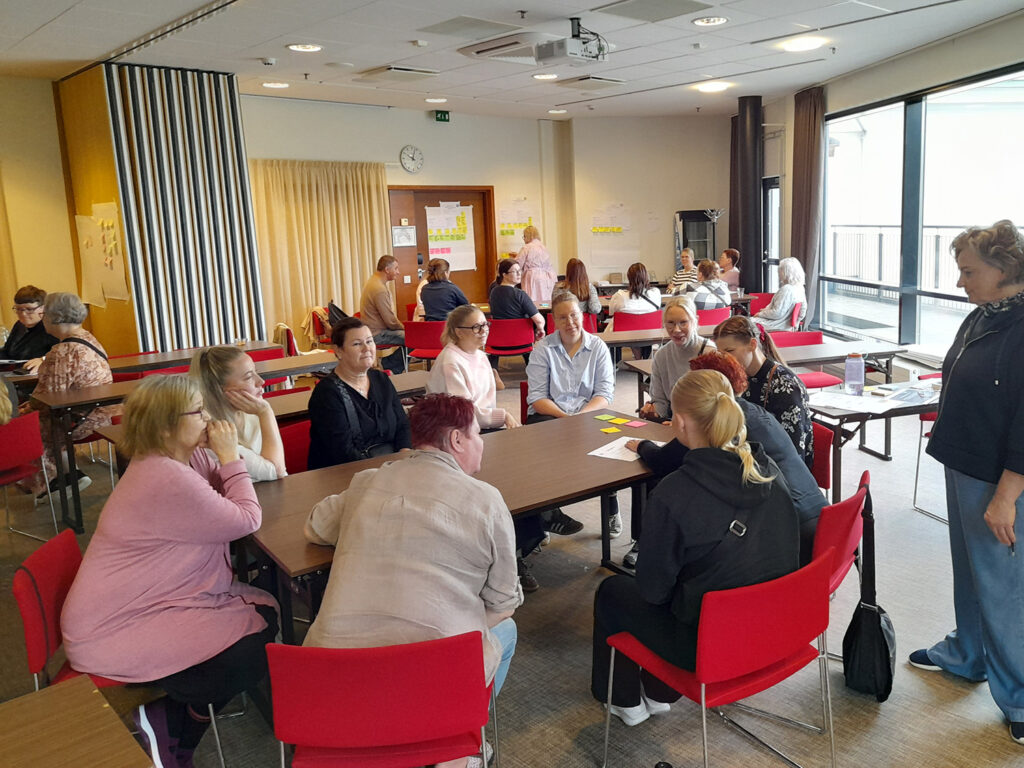 Suuressa kokoustilassa on useampi ryhmä ryhmittyneenä pöytien ympärille keskustelemaan. Oikeassa reunassa näkyy suuret maisemaikkunat ulos.