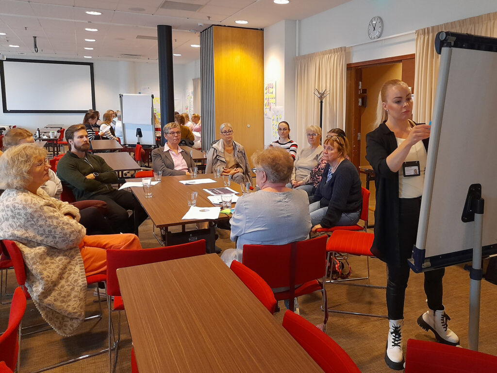 Suuressa kokoustilassa kymmenkunta miestä ja naista istuu pohtimassa ja yksi seisoo ja kirjoittaa fläppitaululle.