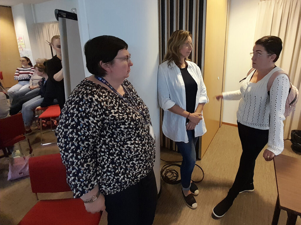 Kolme naista keskustelee seisoen. Kukaan ei katso kameraan. He ovat kokoustilassa ja vasemmassa laidassa näkyy muita ihmisia.