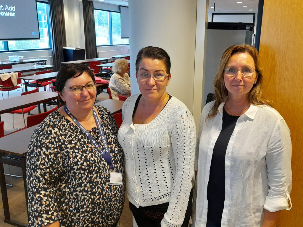 Kameraan katsovat ja hymyilevät kolme naista. Kaikilla on silmälasit. Taustalla suuri kokoustila, jossa näkyy yksi ihminen istumassa ja maisemaan avautuvat isot ikkunat.