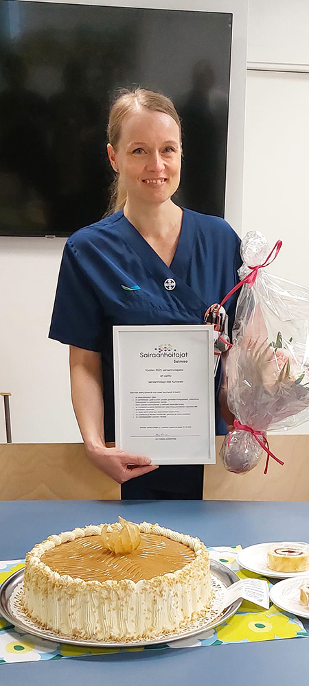 Tummansiniseen hoitaja-asuun pukeutunut nainen hymyilee ja pitää edessään kehystettyä kunniakirjaa ja sellofaaniin pakattua kukkakimppua. Kuvan edustalla on kinuskitäytekakku.