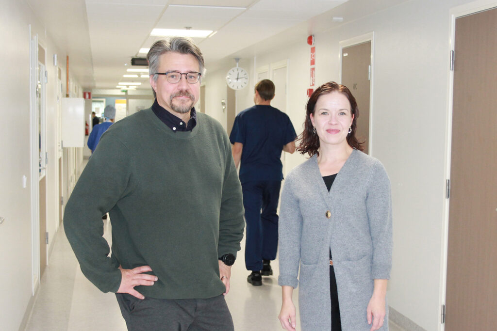 Pitkällä sairaalan käytävällä kuvan etualalla seisovat tummanvihreään villapaitaan pukeutunut harmaantuva nuorekkaan oloinen mies kädet lanteillaan ja harmaaseen pitkään villatakkiin pukeutunut tummahiuksinen nainen. Molemmat hymyilevät. Taustalla kulkee hoitohenkilöstöä hoitajan asuissaan. 