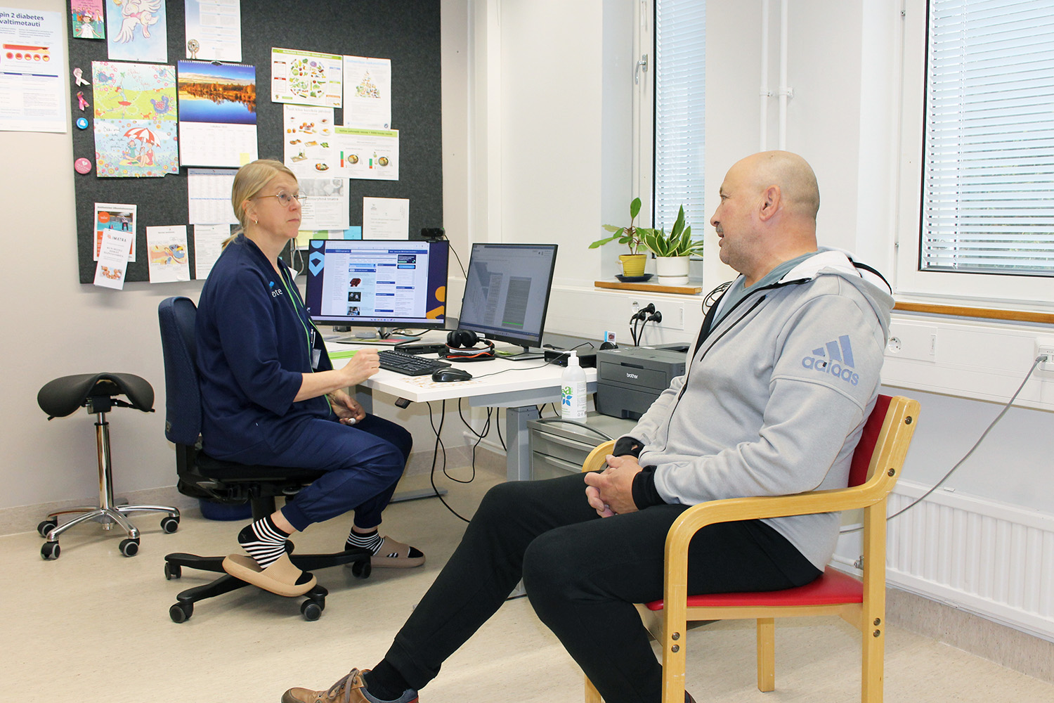 Vastaanottohuoneessa kaksi henkilöä istuu keskustelemassa. Toinen henkilö istuu punaisella tuolilla harmaassa Adidas-hupparissa, toinen työtuolilla sinisessä työasussa tietokoneen ääressä. Työpöydällä on kaksi näyttöä, näppäimistö ja kasvi ikkunalaudalla. Taustalla ilmoitustaulu, jossa on värikkäitä piirustuksia ja papereita.