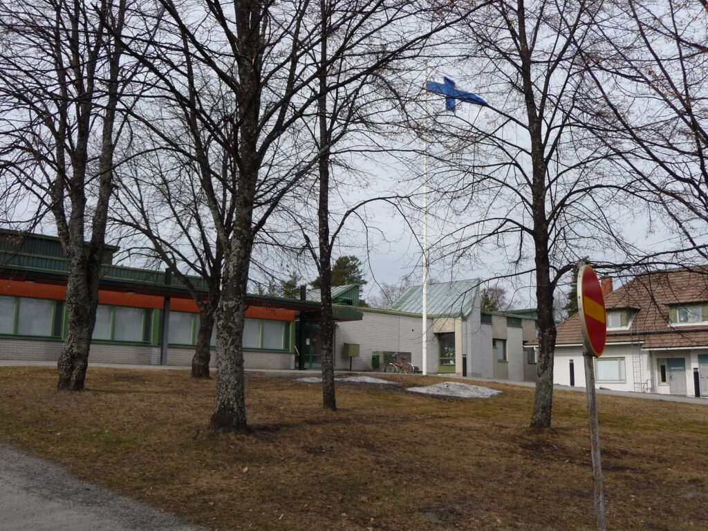 Etualalla olevien lehdettömien puiden rankojen takaa näkyy matala rakennus, jossa tiilenpunaista väriä. Puiden keskellä pihassa on myös Suomen lippu liehumassa lipputangossa. 