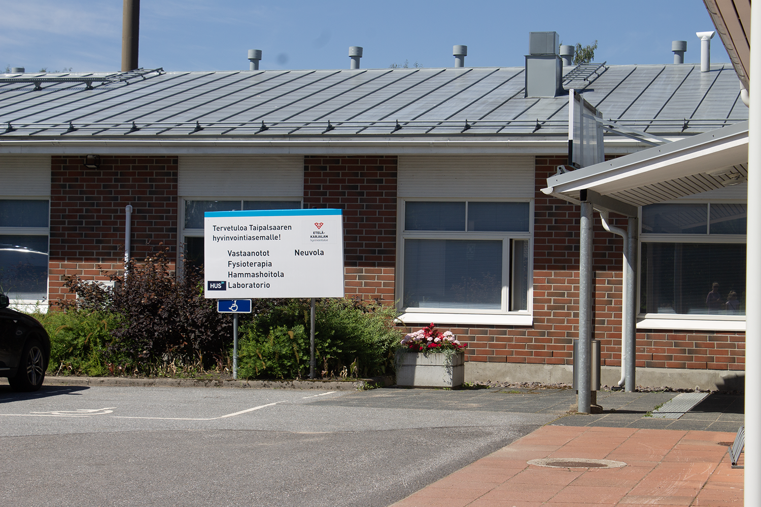 Lähikuva Taipalsaaren hyvinvointiaseman sisäänkäynnistä. Edessä näkyy asfaltoitua pihaa, kyltti ja ikkunoita. Rakennus on punatiilinen matala rakennus, jossa valkoiset ikkunat.