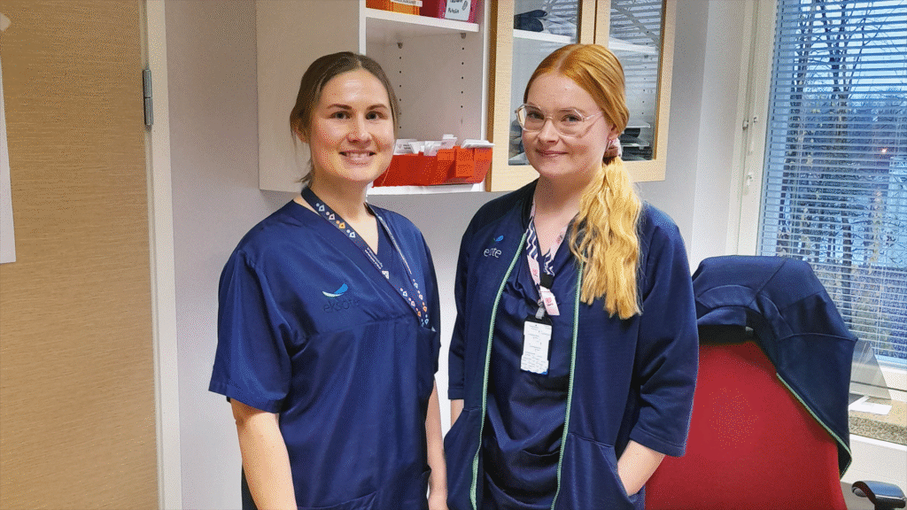 Kaksi hiukset taakse sitonutta tummansinisiin hoitajan asuihin pukeutunutta naista seisoo hymyillen hoitohuoneessa.