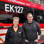 Hymyilevät nainen ja mies seisovat hälytysajoneuvon edessä. 