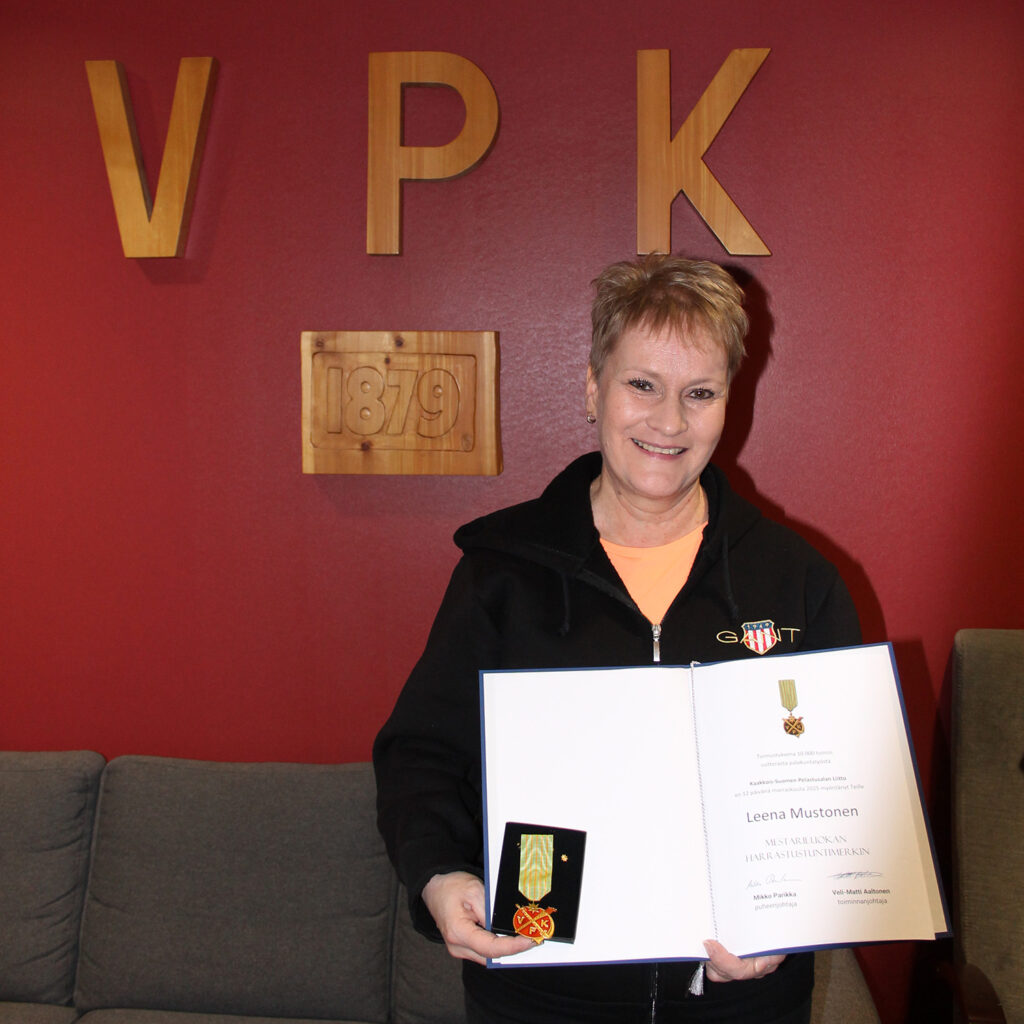 Hymyilevä nainen pitelee käsissään kunniakirjaa ja kunniamerkkiä. Taustalla tummanpunainen seinä, jolla oranssit kirjaimet V, P ja K.
