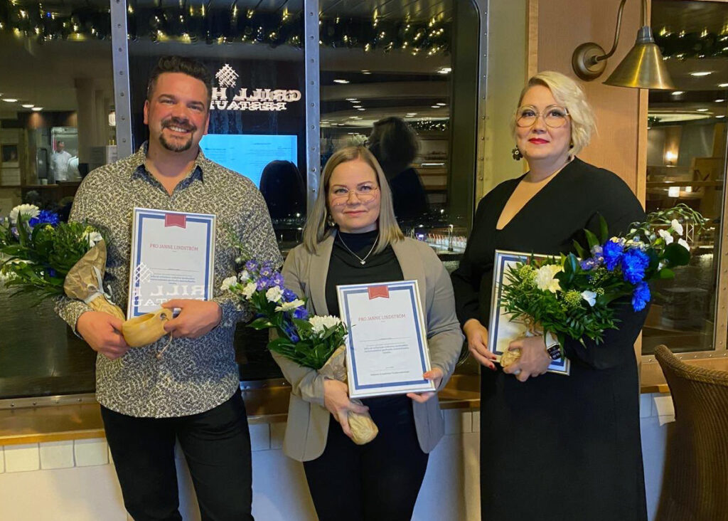 Kolme henkilöä seisoo sisätiloissa suuren ikkunan edessä, jossa näkyy iltavalaistus ja heijastuksia ulkoa. Jokaisella on kädessään kukkakimppu, jossa on valkoisia ja sinisiä kukkia sekä vihreitä lehtiä, ja lisäksi kehystetty kunniakirja. Taustalla näkyy ravintolamainen ympäristö, lämmin valaistus ja koristevaloja ikkunan yläreunassa.