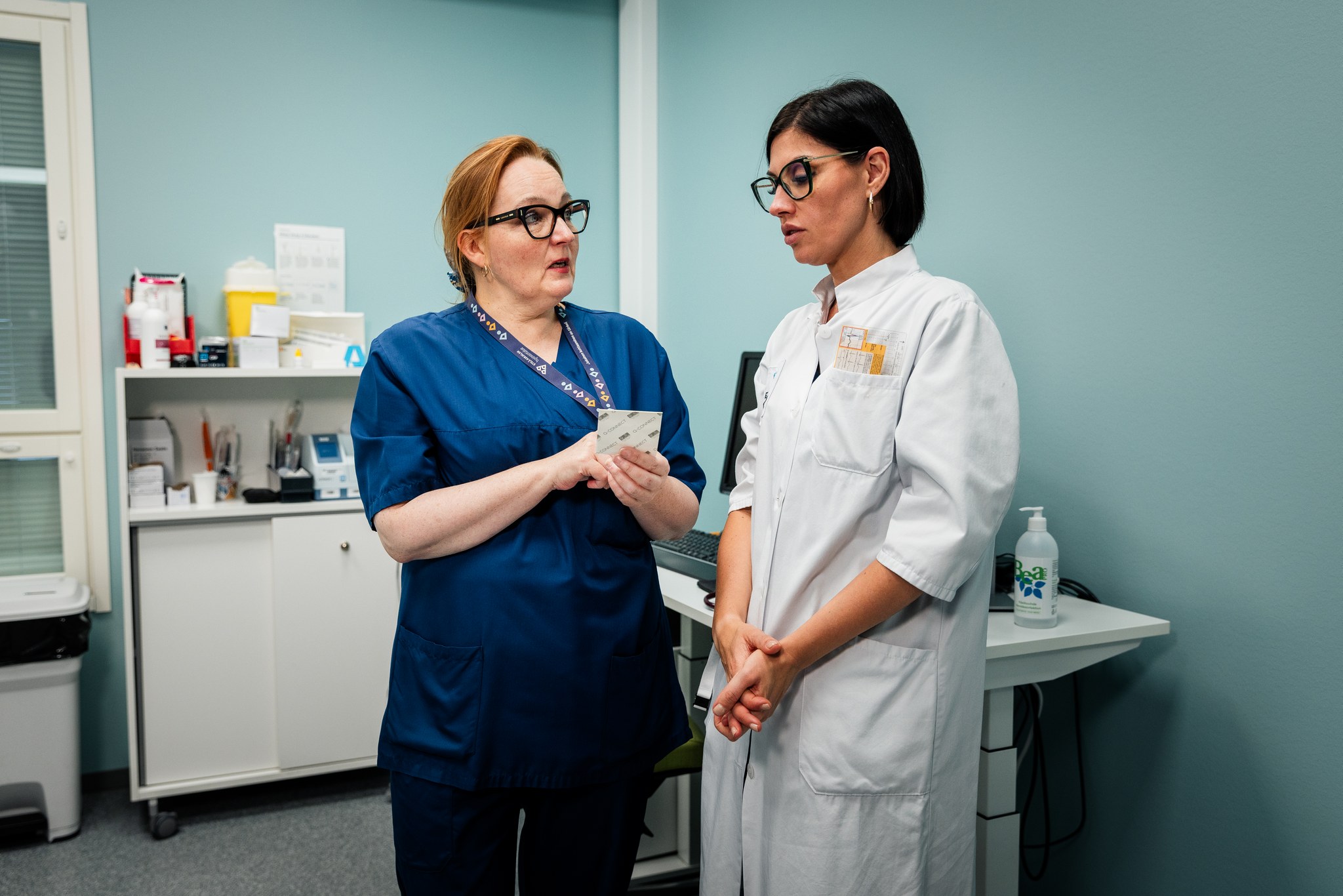 Edestäpäin kuvatut kaksi seisovaa naista. Vaatteiden perusteella he ovat sairaanhoitaja ja lääkäri. Sairaanhoitaja osoittaa toisella kädellään kädessään pitelemäänsä muistilappua ja kysyy lääkäriltä jotain. Taustalla on vastaanottohuoneen tarpeistoa ja vaalean sinivihreä seinä.